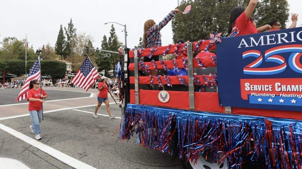 A patriotic parade float adorned in red, white, and blue travels down a Mission Viejo street, greeted by enthusiastic spectators and waving American flags during the America 250 Parade. The community gathers to celebrate and recognize Service Champions—your trusted local experts dedicated to providing reliable HVAC, plumbing, and drain services with genuine care for Southern California families’ home comfort needs.