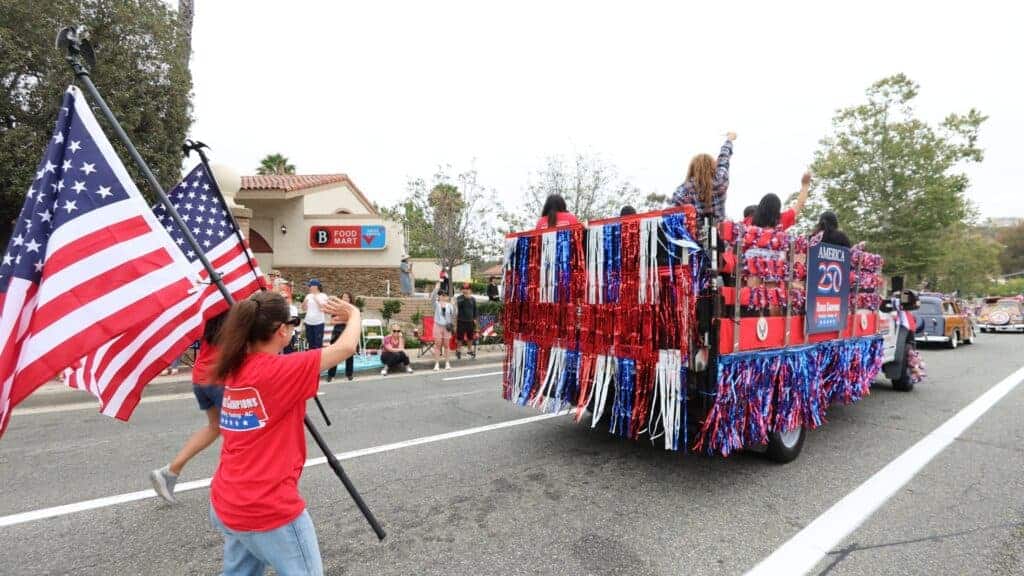 A welcoming woman waves an American flag near a festive parade float adorned with red, white, and blue streamers and community members riding along, all traveling down the street during the Mission Viejo America 250 Parade—a celebration that highlights local spirit and togetherness, values shared by Service Champions as your trusted partner in home comfort services throughout Southern California.