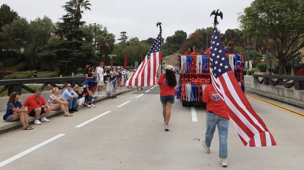 Service Champions team members and local spectators watch supportively from the roadside as two individuals carry large American flags behind a festively decorated float during the America 250 Parade in Mission Viejo, proudly highlighting Service Champions’ role as Southern California’s trusted partner for home comfort, HVAC, plumbing, and drain services.