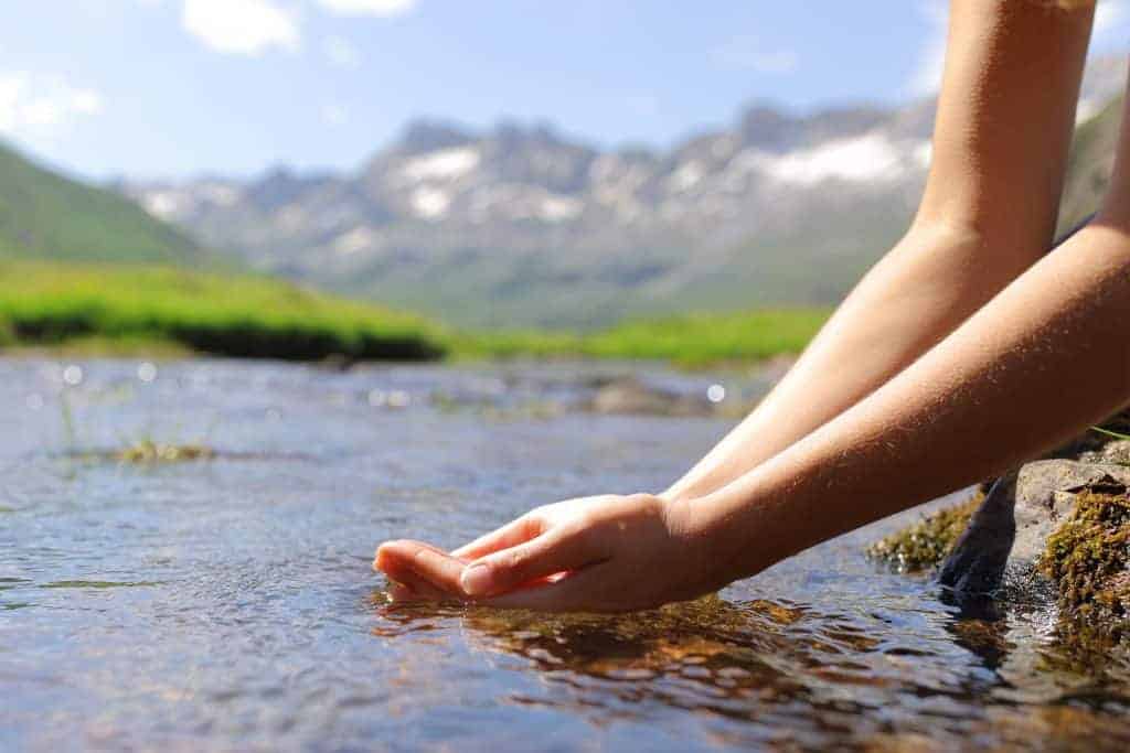 Hands touching flowing river water in scenic mountain landscape.