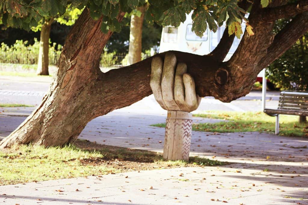 A large, falling tree trunk in a park is held up by a strong, intricately carved hand made of wood.