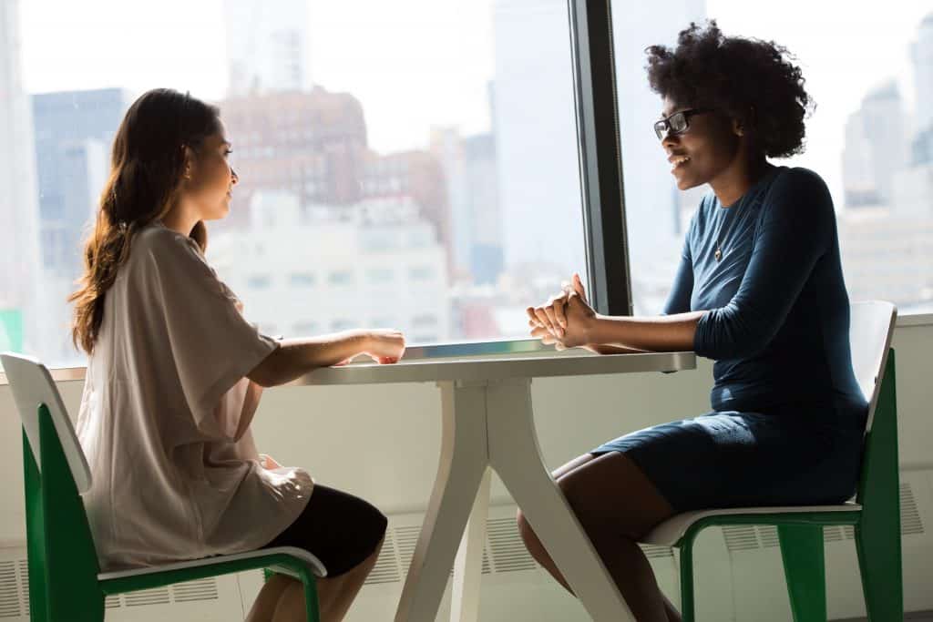 Two women sit across from each other at a table in front of a window, deep in conversation.