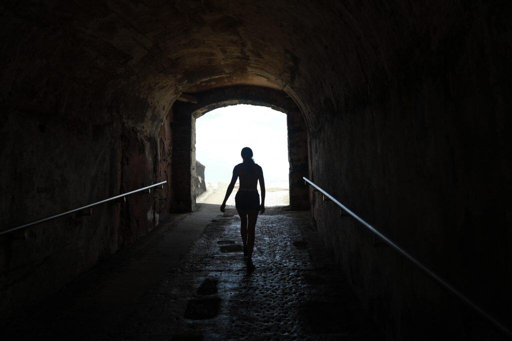 A woman walks through a dark tunnel towards and opening of light, her shape silhouetted by the bright natural light.