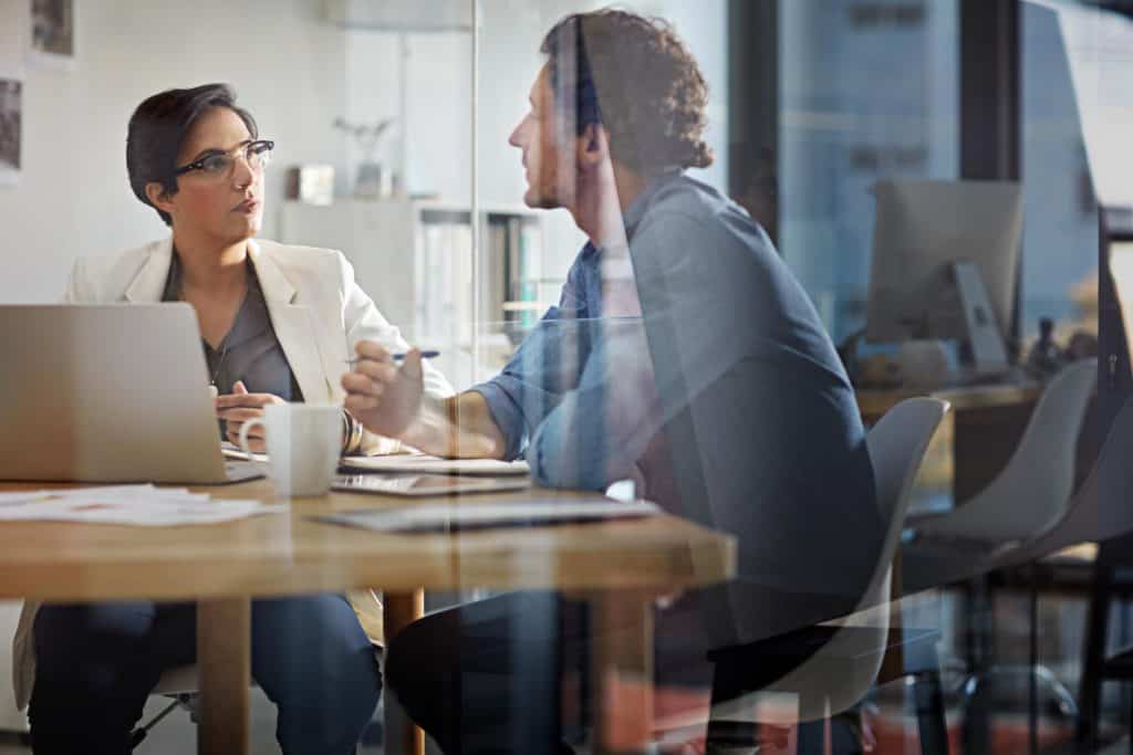 A man and a woman sit at a desk behind a glass wall, having a discussion. The woman listens to the man talking. There's paperwork, laptops, and coffee cups on the table in front of them.