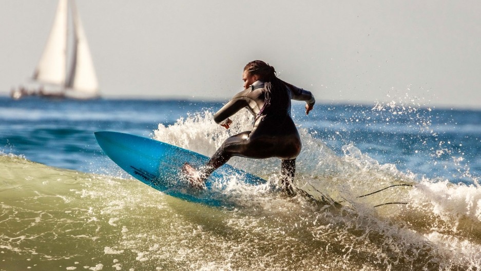 A male surfer rides a capping wave in the ocean. His back is to the camera as he balances on a blue surf board. There's a sail boat in the background.