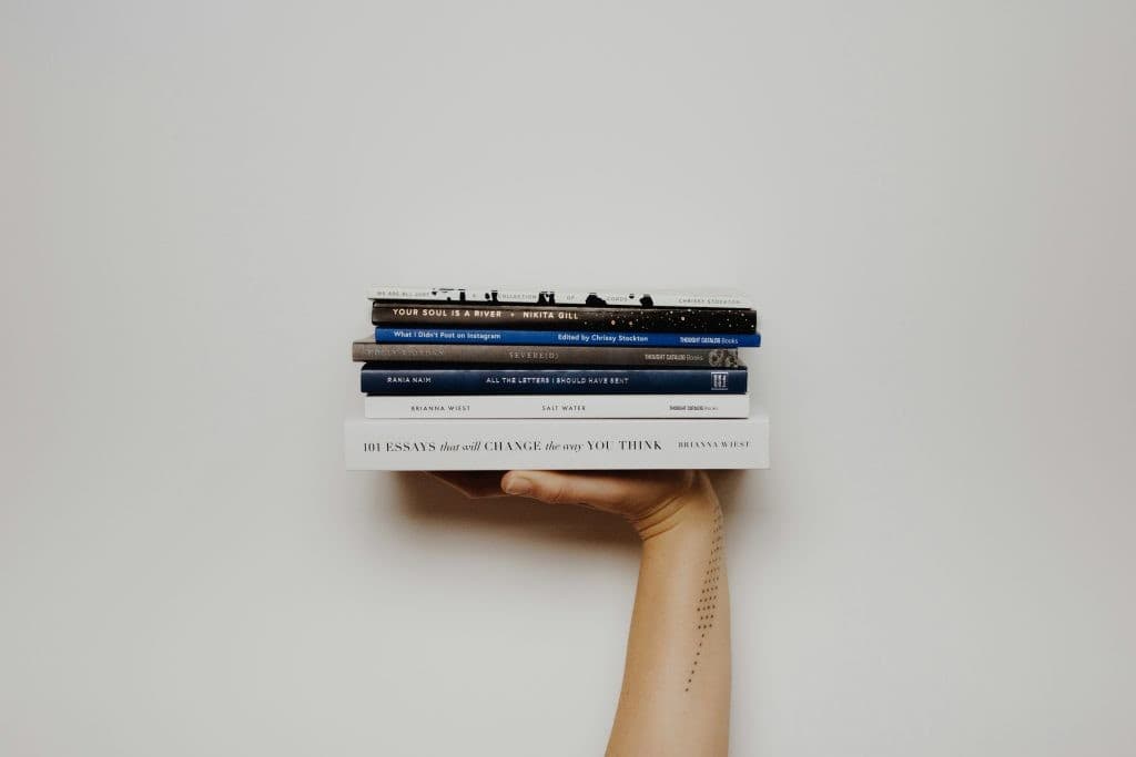 A person holds a stack of 7 books in their hand against a white wall.