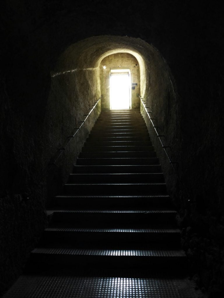 A photo shows a view point of a dark staircase looking up, towards a light-filled arched doorway at the top of the stairs.