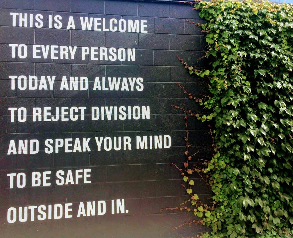 A sign with greenery to the side that says "This is a welcome to every person today and always to reject division and speak your mind to be safe outside and in"