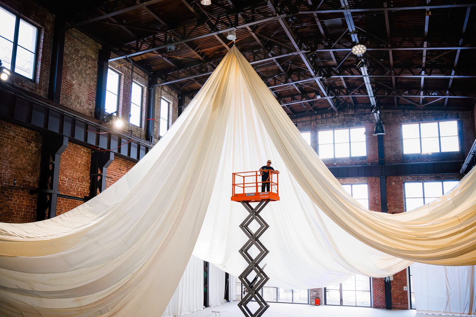Ivory drapery being rigged high in a daylight-filled industrial hall, with an installer on a lift showing the scale
