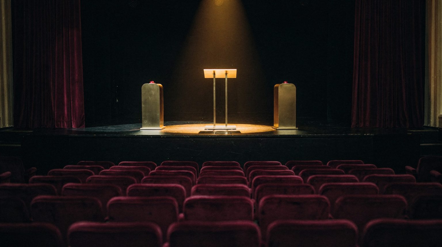 An empty theatre seen from behind rows of deep plum velvet seats, looking at a low stage where a single warm spotlight falls on a slim lectern flanked by two brushed-brass buzzer podiums, with heavy dark red curtains at the wings.