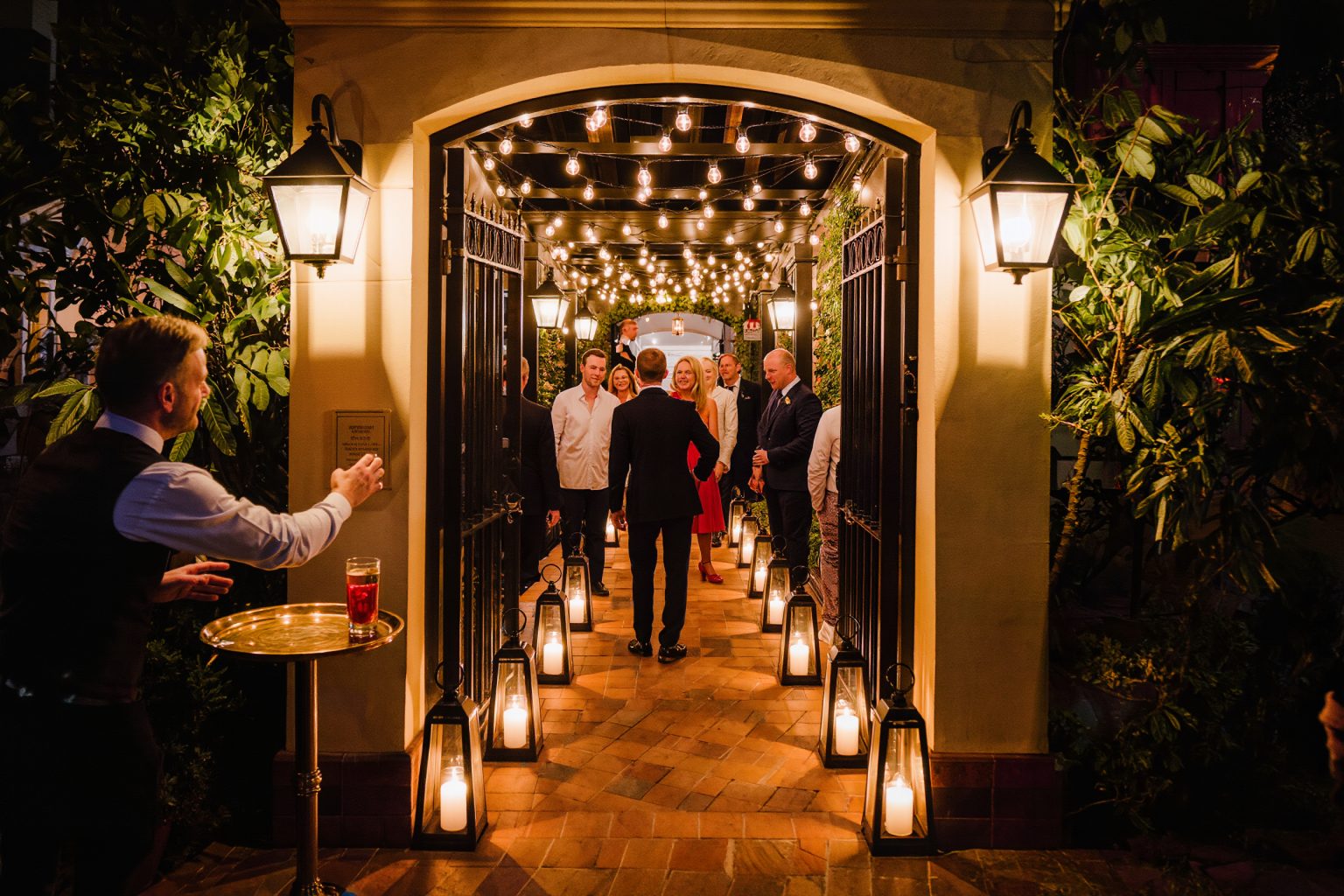 Guests stepping through a warmly lit garden gate where a greeter offers welcome drinks