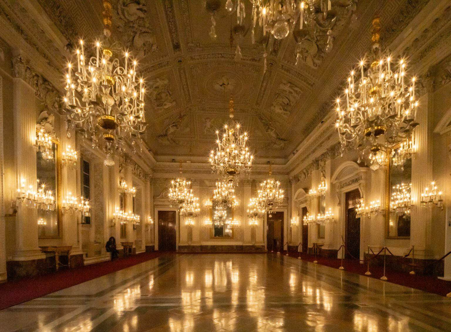 An ornate ballroom features large crystal chandeliers hanging above a polished floor.