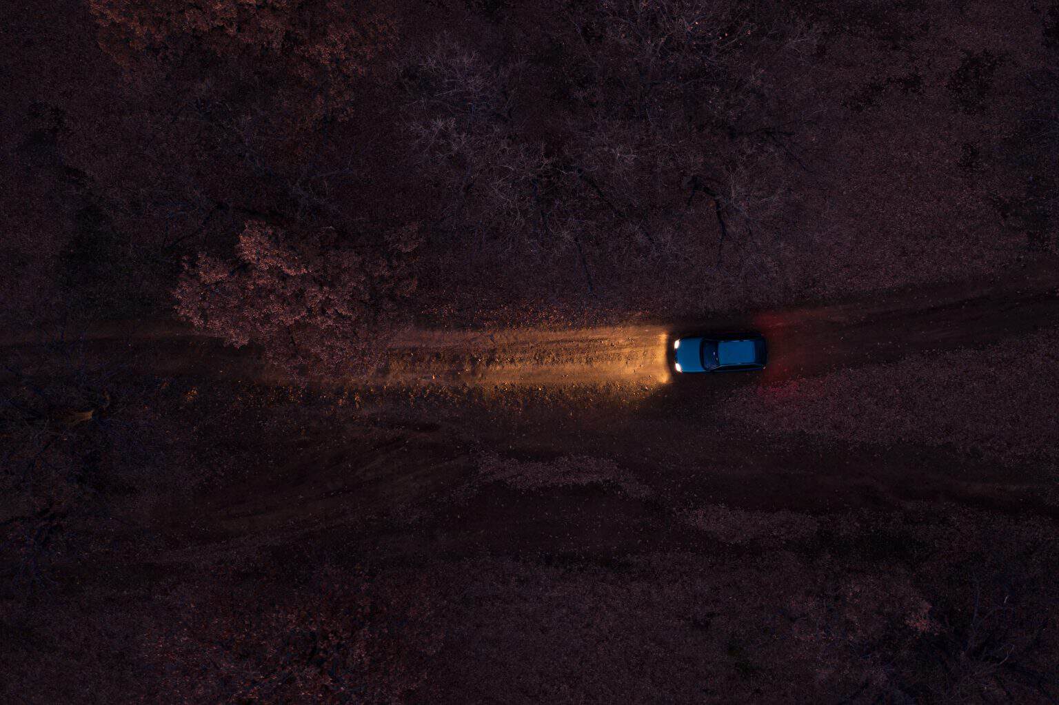 Seen from directly overhead, a single car throws a cone of headlight along an unlit dirt track through dark woodland