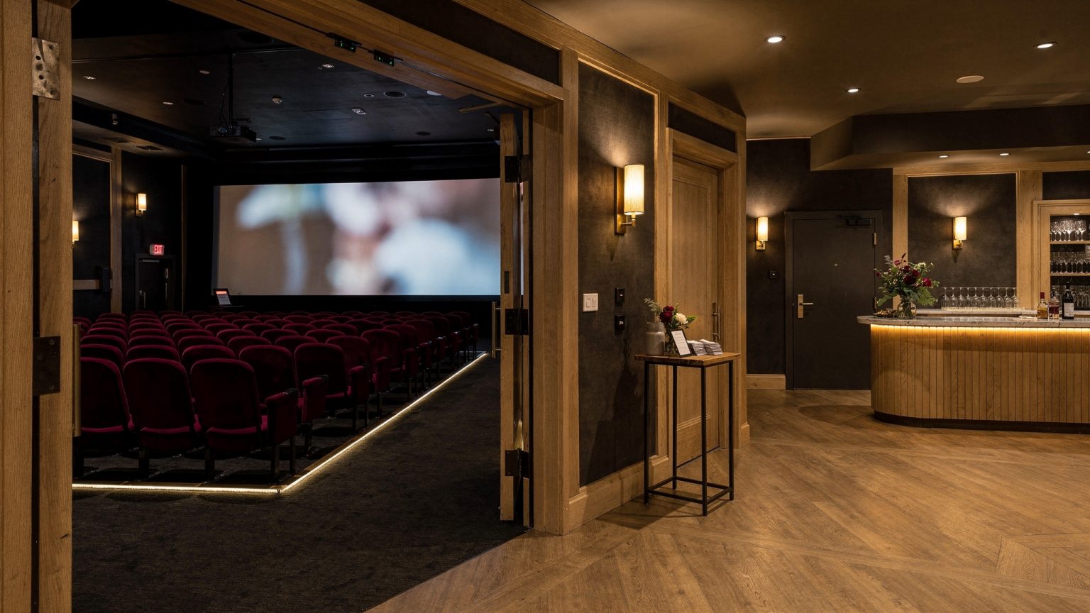 Independent cinema auditorium viewed from the lobby threshold with clear aisle lighting, a glowing screen and a compact event welcome and refreshment area.