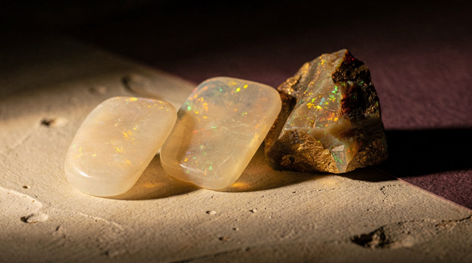 A close study of two polished milky opal slabs and one rough uncut opal resting on a bone-coloured plaster ledge, low raking light igniting green and gold fire inside the stone, against a deep plum shadow.