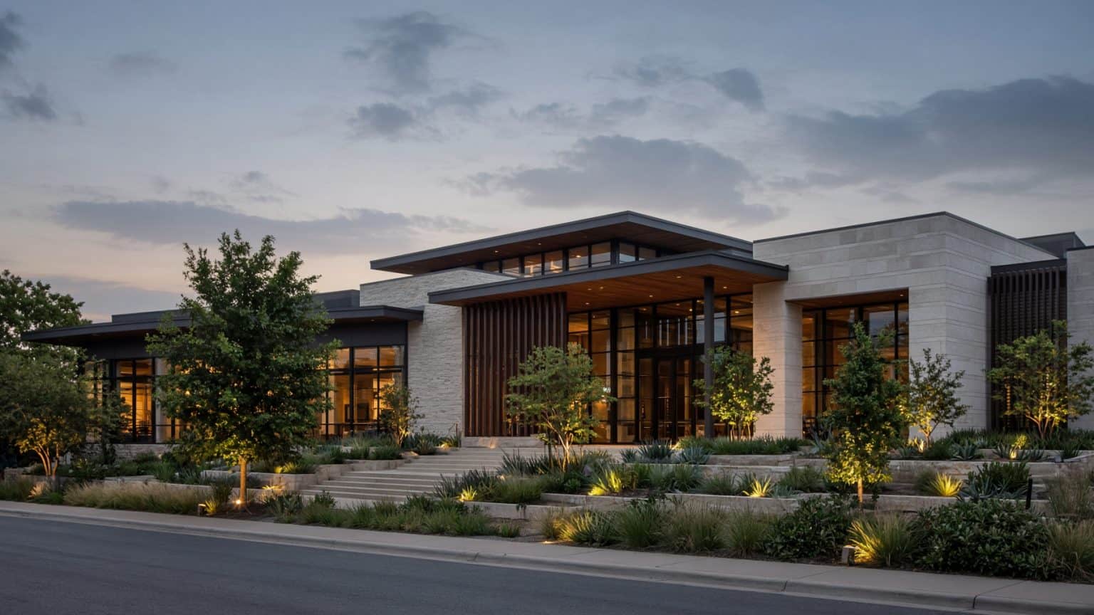 Contemporary multi-level stone and glass event center with warm interior lighting, landscaped stairs and desert-style plantings at twilight