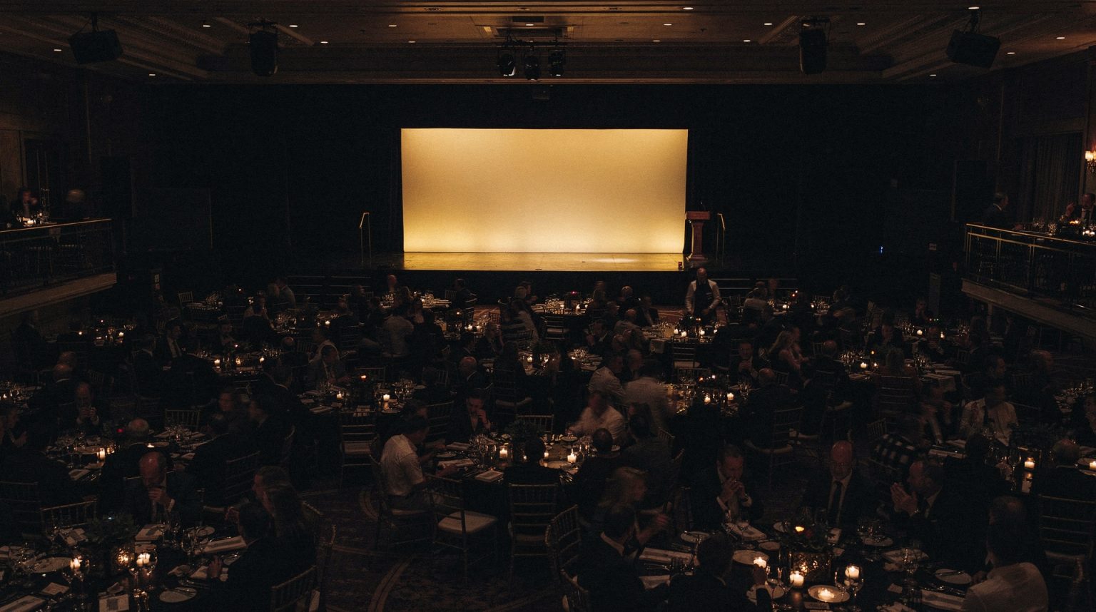 A large gala dining room seen from a dark balcony at the back: a blank warmly lit stage and lectern at the far end, and between it and the camera an unlit sea of round tables and seated guests reduced to dark shapes and points of candlelight.