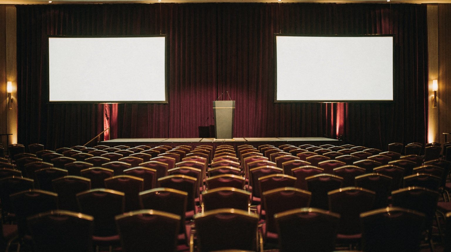 A large hall set theatre style: long rows of deep plum banqueting chairs facing a low stage with a slim lectern, flanked by two wide projection screens glowing an even blank white against heavy dark red drapes.