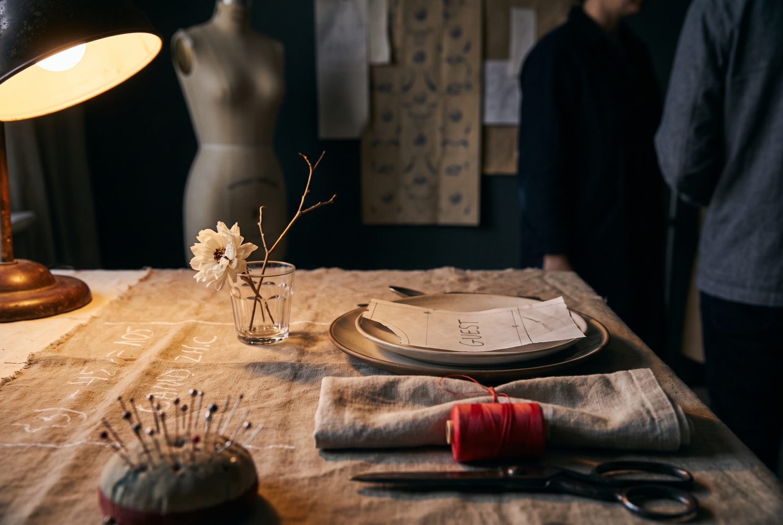 A pin cushion, tailor's shears and a spool of red thread sit beside a single dried bloom on a raw, pin-tacked calico tablecloth marked in chalk.