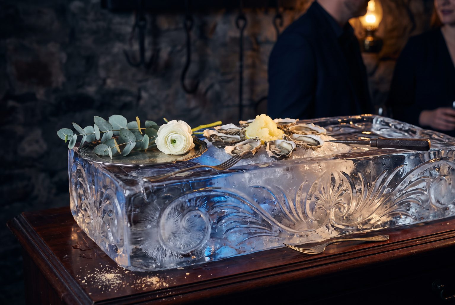 Oysters and granita set directly into a carved ice trough catch warm gaslight beside a sprig of silver eucalyptus and a tarnished silver oyster fork.
