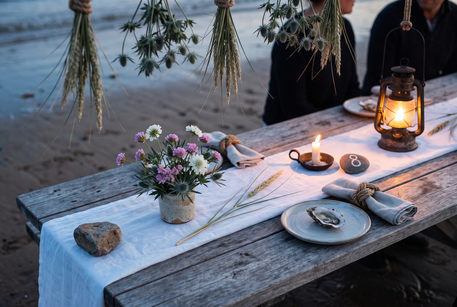 A salt-glazed stoneware cup of sea holly, thrift and white scabious sits beside an oyster-shell place setting on a weathered driftwood table.