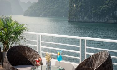 Luxury cruise deck with scenic bay view, wicker chairs, tropical drinks, and mountain landscape.