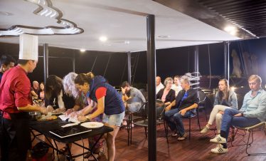 Cooking class on a boat with chefs and tourists enjoying a Vietnamese culinary experience at night.