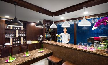 Elegant hotel bar with bartender pouring wine, luxurious wooden furnishings, and vibrant orchids.