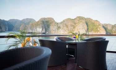 Panoramic view of a luxury cruise deck with wicker chairs and tropical plants, overlooking stunning limestone islands in Halong Bay.