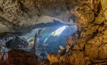 Imposing cave with sunlight rays, limestone formations, and tourists exploring inside.