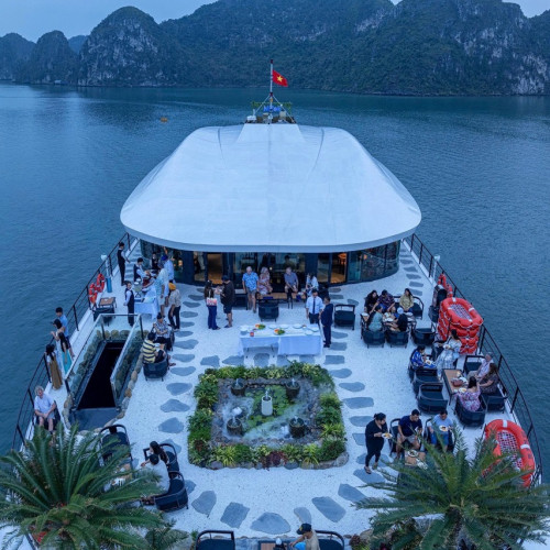 Aerial view of a restaurant boat with groups of people dining on the deck, surrounded by water and rocky islands; a small garden centerpiece is in the middle.