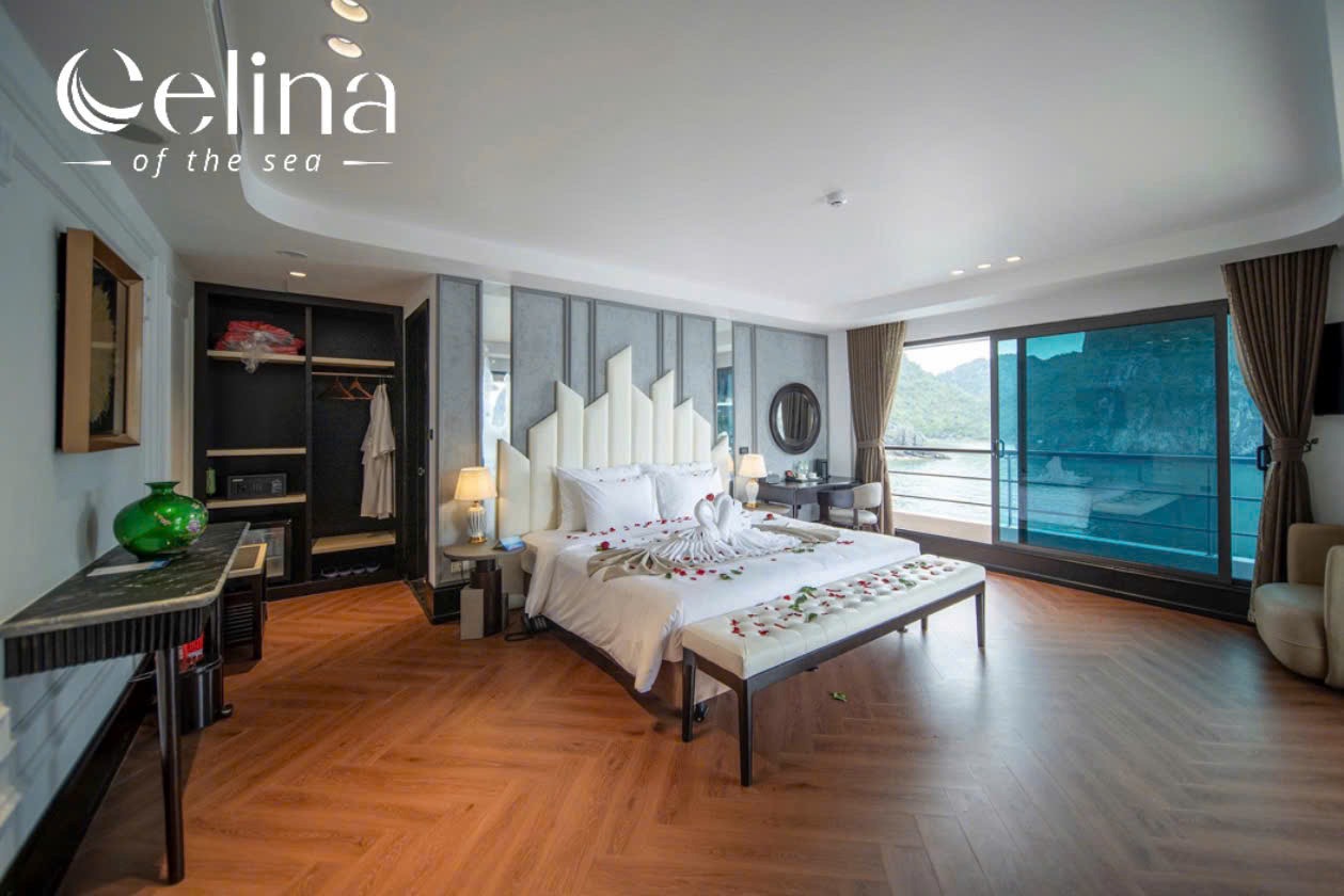 Luxury hotel bedroom with a bed dressed in white, a towel swan centerpiece and rose petals, bench at foot, and a balcony with sea views.