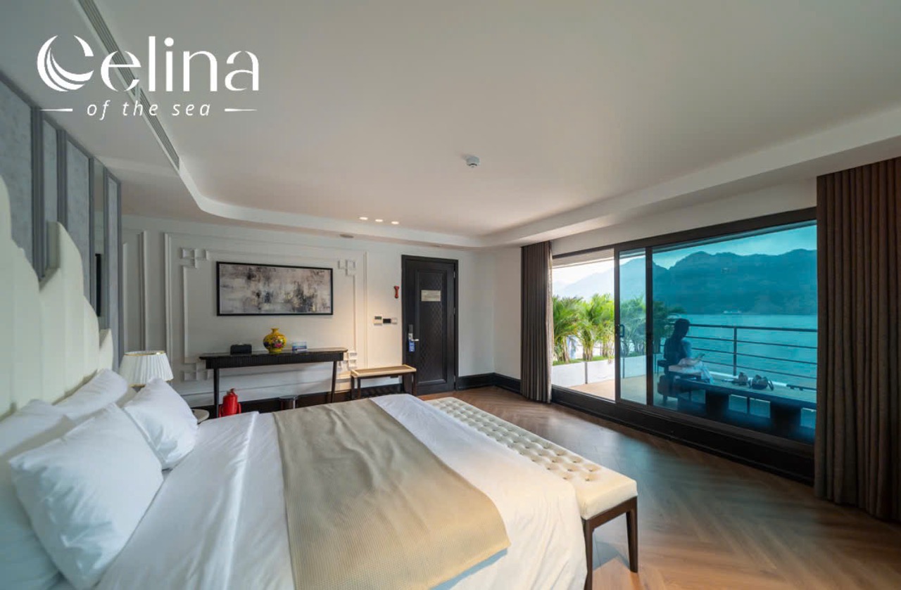 Hotel bedroom with a large bed, bench at the foot, and sliding doors to a balcony overlooking the sea and mountains.
