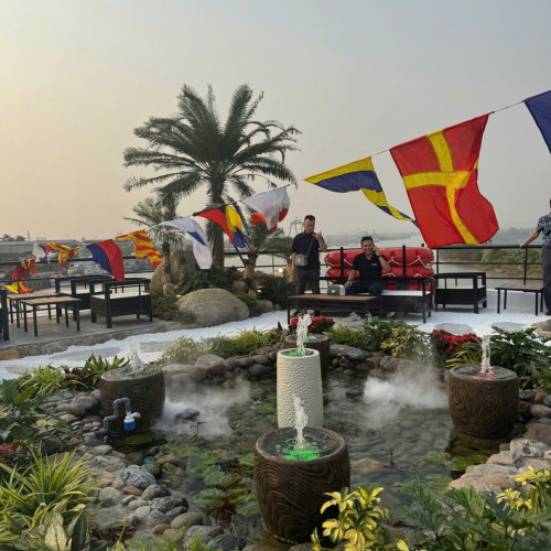 Rooftop garden with decorative fountains, rocks, and lush plants, set against colorful pennant flags and a seating area in the background.