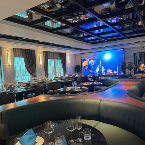 Upscale banquet room with curved leather booths, round tables set with blue napkins and glassware, and a large screen at the front for presentations/entertainment.