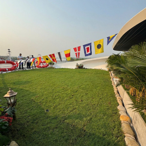 People stand along a line of colorful signal flags strung above a grassy terrace, with a curved white building and tropical plants on the right and water in the distance.
