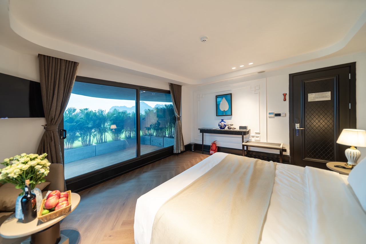 King bed in a bright hotel room with sliding glass doors to a balcony and palm-filled outdoor view on a sunny day.