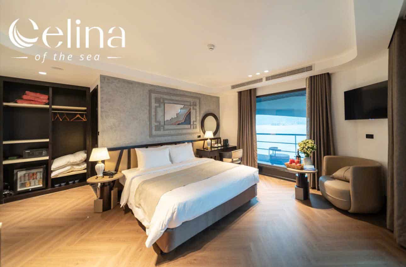 Elina of the sea watermark in the top-left corner of a luxurious hotel room with a king bed, nightstands, open wardrobe, and balcony overlooking water
