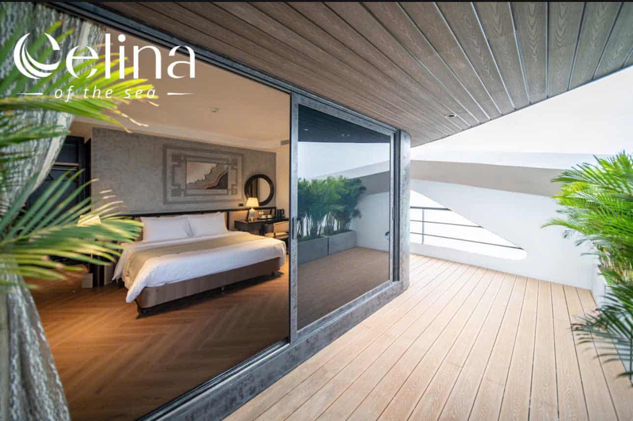 Hotel bedroom with a sliding glass door opening to a wooden balcony, tropical plants outside, and the 'Elina of the sea' logo in the corner.