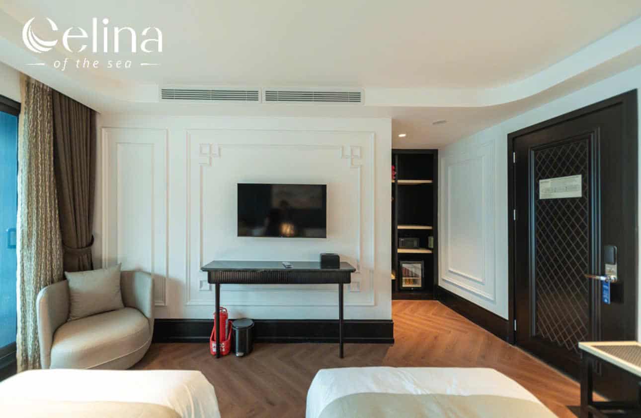Hotel room with wall-mounted TV above a black console, beige seating chair, and two beds at the foreground; dark door on the right and built-in shelves near the entry. Logo 'Celina of the sea' at top left.