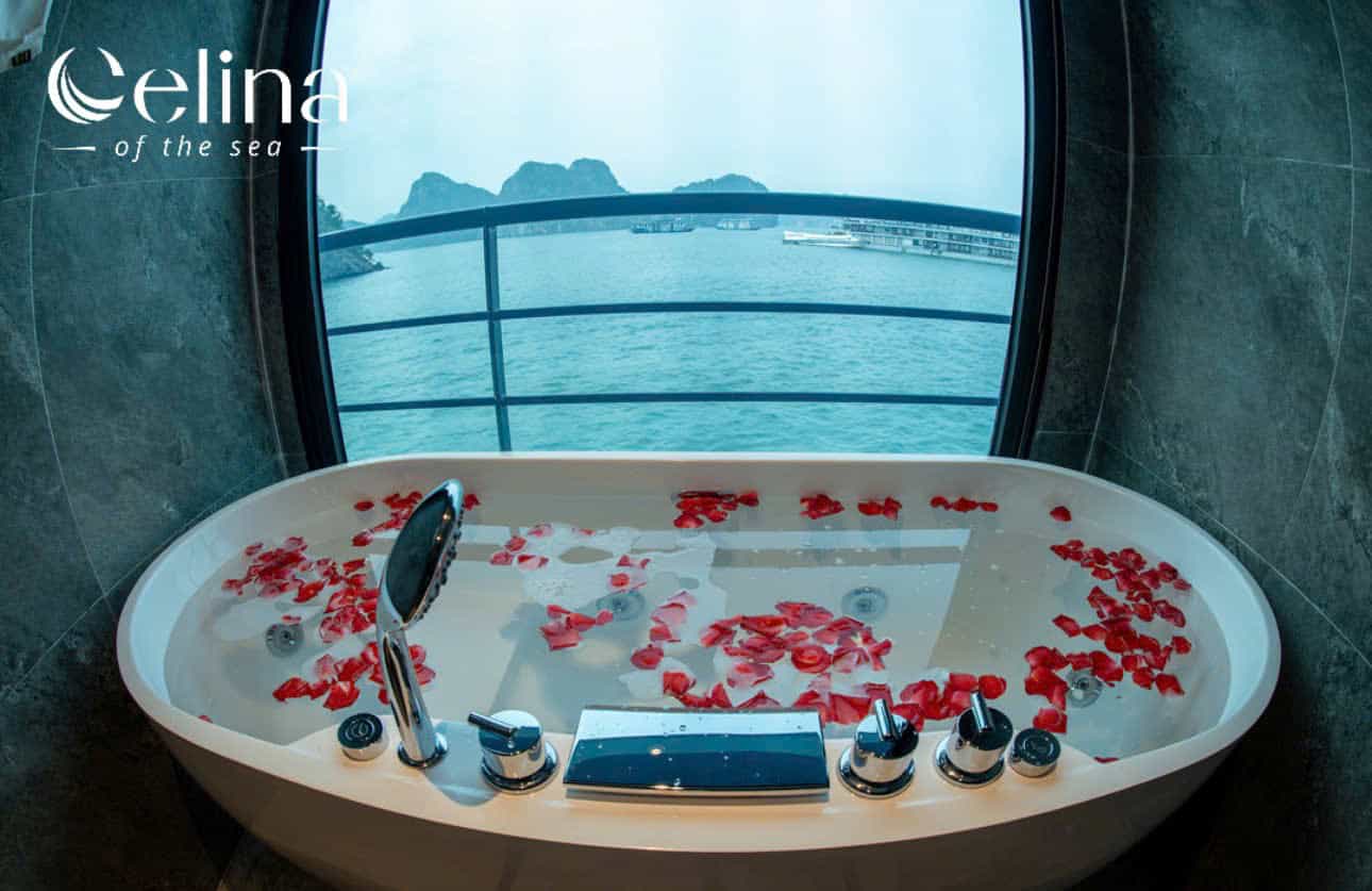 Luxurious white bathtub filled with water and red rose petals, ocean view through a curved window; Celina logo in the top-left corner.
