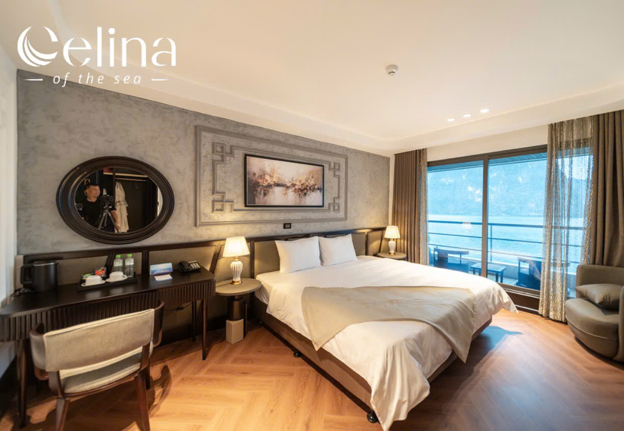 Hotel bedroom with a king bed, two nightstands with lamps, a dark wood desk, and a balcony door showing an ocean view; Celina of the sea branding in the top-left corner.