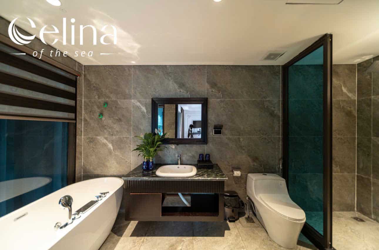 Luxurious marble bathroom with a freestanding bathtub on the left, a dark wood vanity with sink and mirror in the center, and a glass-enclosed shower on the right. A potted plant and towels sit on the vanity area.