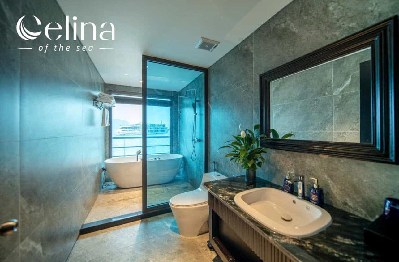 Luxurious bathroom with gray marble walls, a marble countertop sink, and a large black-framed mirror; a glass partition reveals a freestanding tub and an ocean view beyond the balcony.