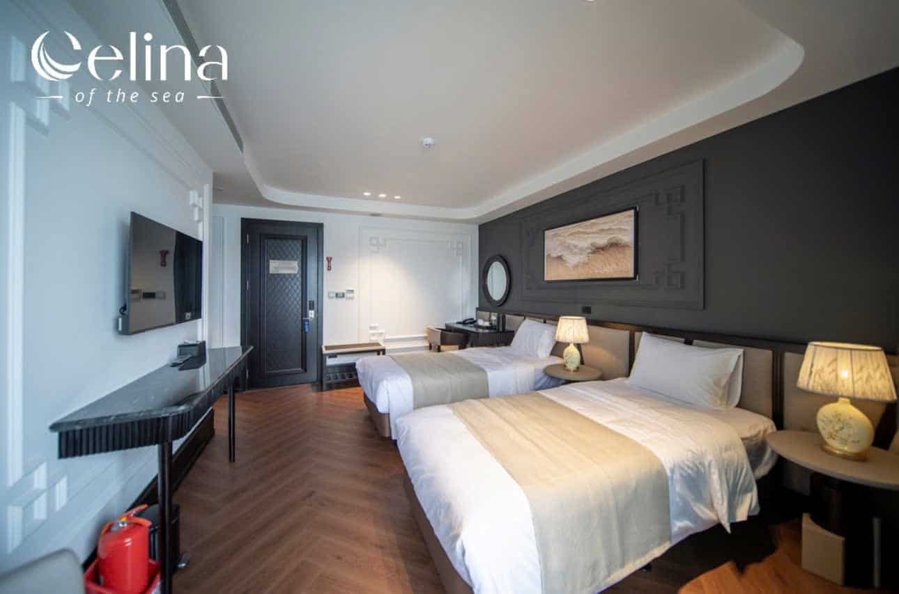 Hotel room with two tidy beds, beige bed runners, and warm lamps on round side tables; dark accent wall and framed art above the beds.