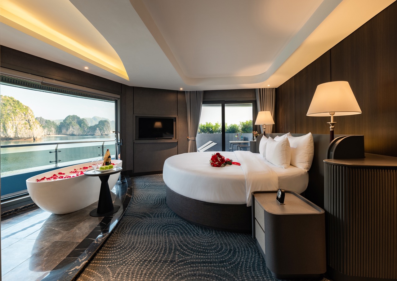 Luxurious hotel suite with a round bed, large windows, and a balcony overlooking a blue bay. A freestanding bathtub with rose petals sits nearby, creating a romantic ambience.