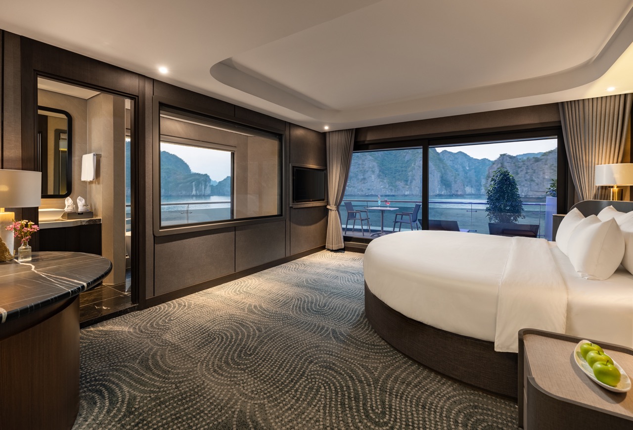 Luxurious hotel suite with a round bed, dark wood accents, and a balcony overlooking a sea inlet and cliffs.