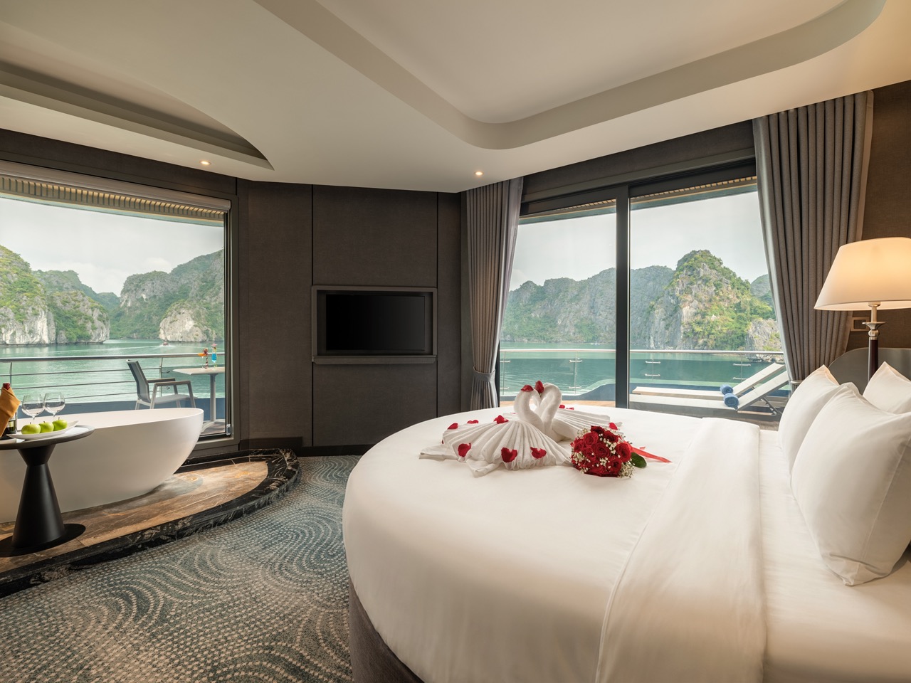Luxurious hotel room with a king bed, towel swans and red bouquet, overlooking a bay through glass doors.