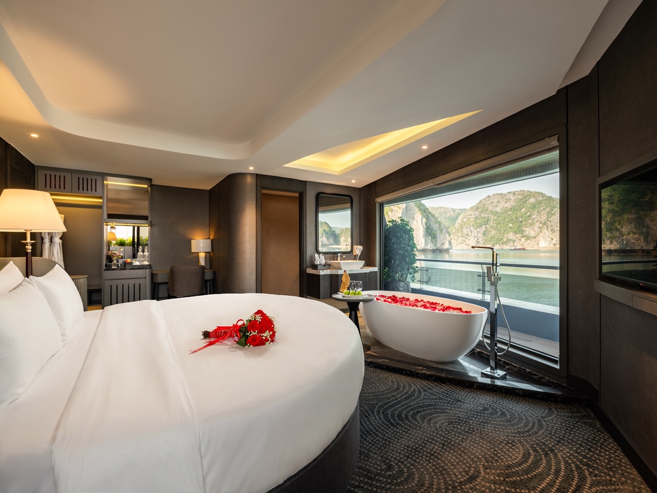 Luxury hotel suite with a large white bed and a bouquet of red roses, plus a freestanding tub by a window overlooking a rocky coastline and water.