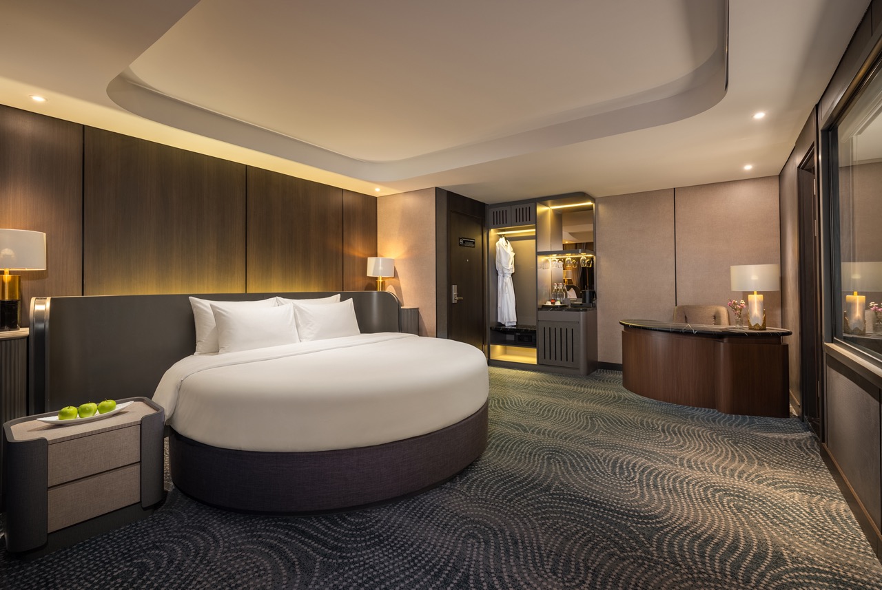 Luxurious hotel room with a circular bed, warm ambient lighting, and soft white bedding on a dark upholstered base, side tables on either side, and lamps.